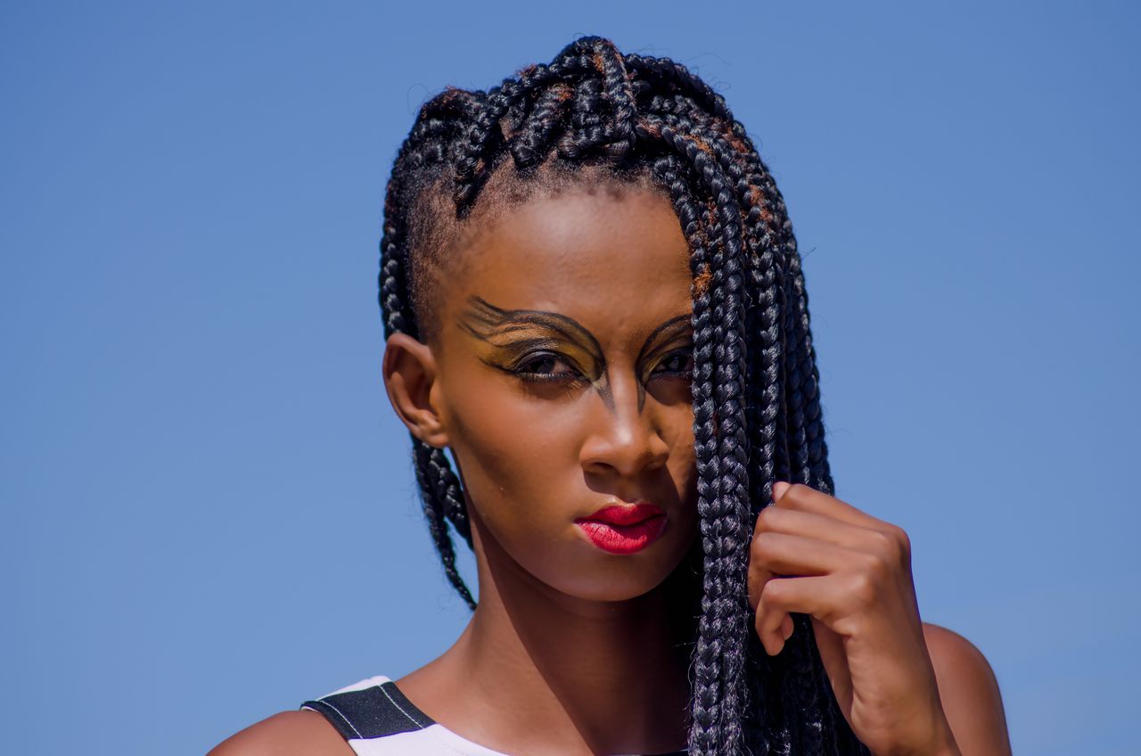 Home Striking portrait of a woman with bold makeup and intricate braids, set against a clear blue sky.