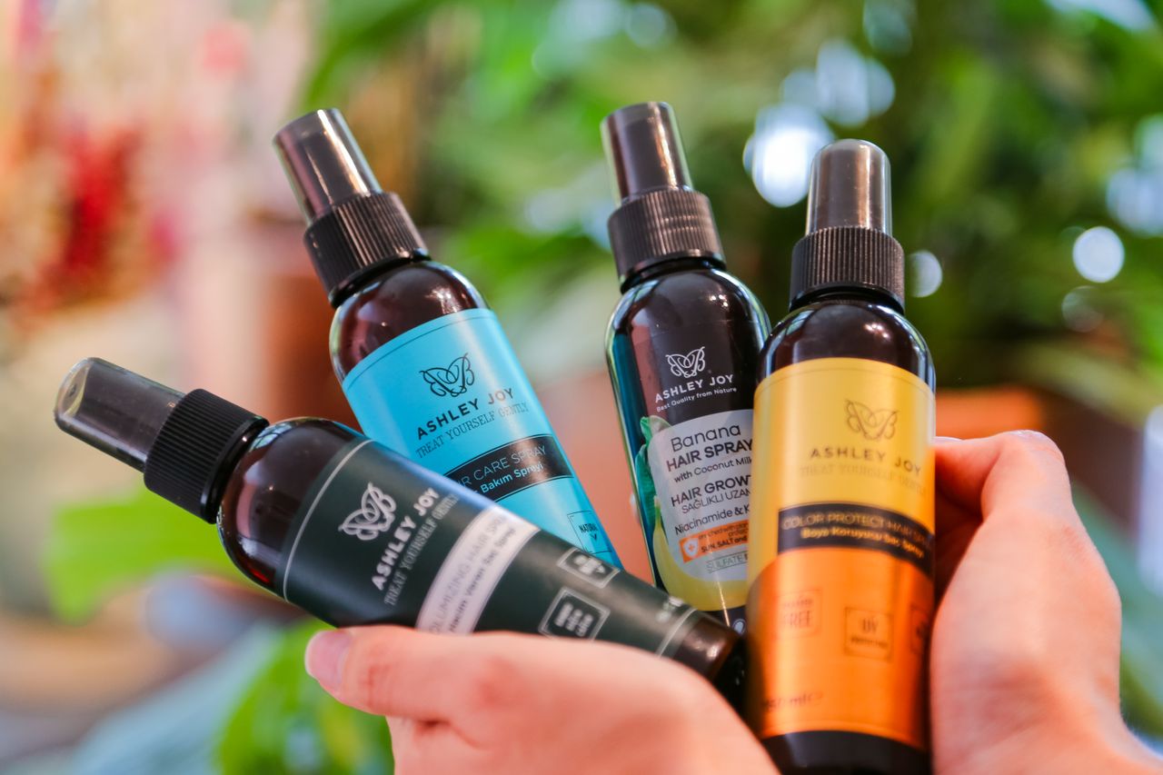 Home Close-up of hands holding various Ashley Joy hair care bottles with a blurred natural background.