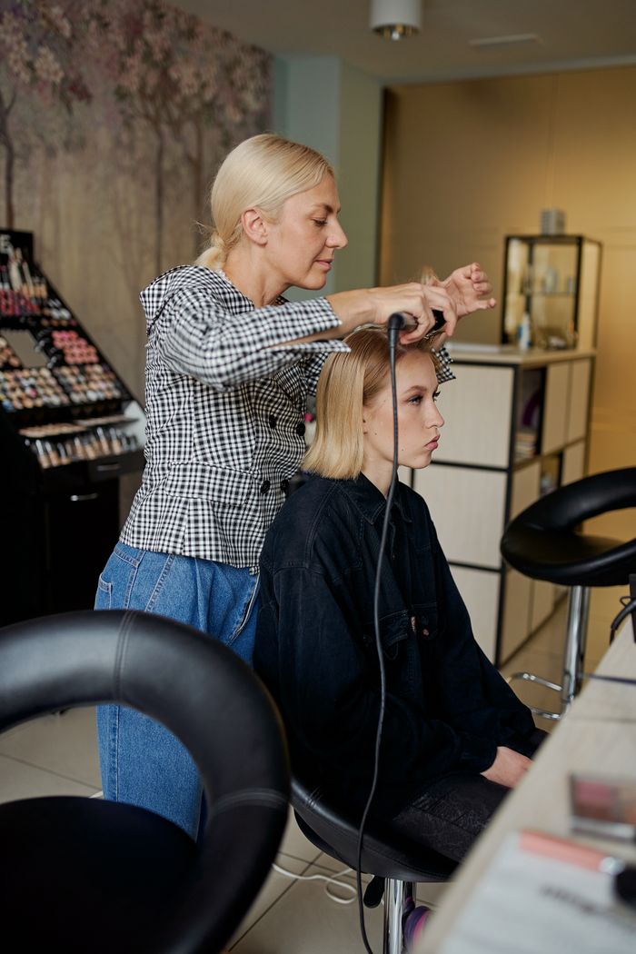 Home Side view of focused hairdresser straightening hair of confident female client with iron in hairdressing salon