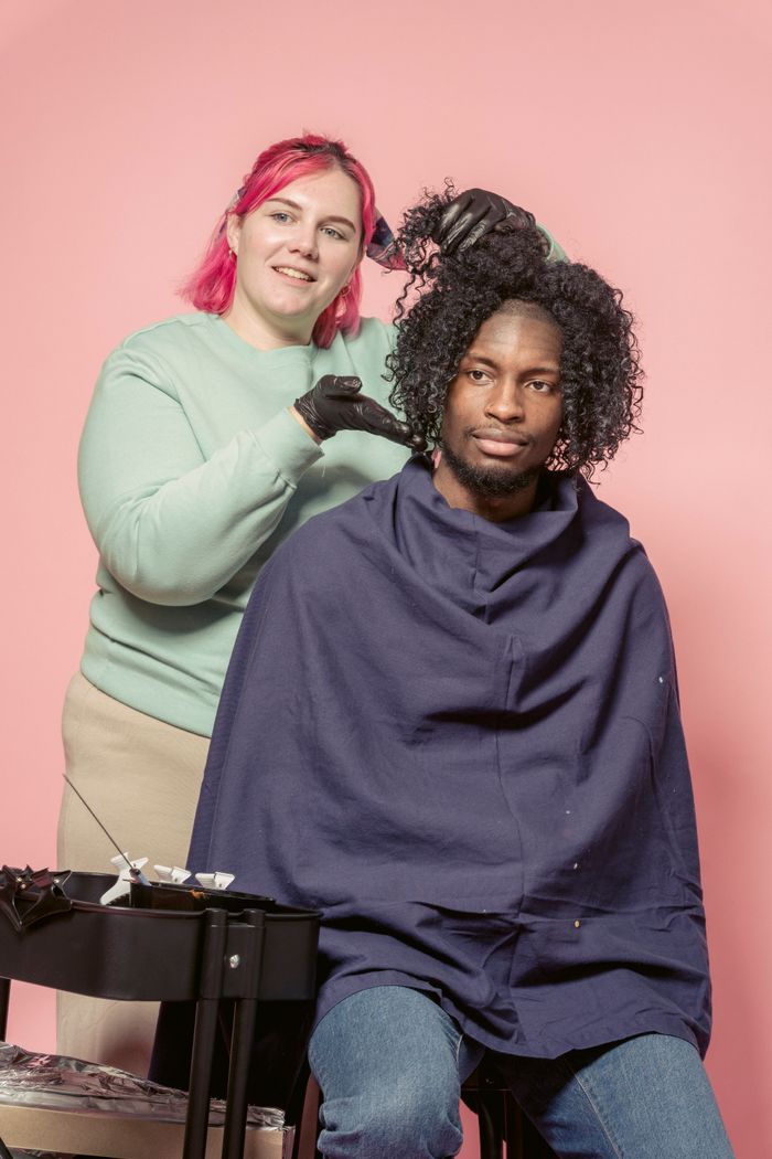 Home Professional female hairdresser in gloves standing African American customer sitting on chair against pink background