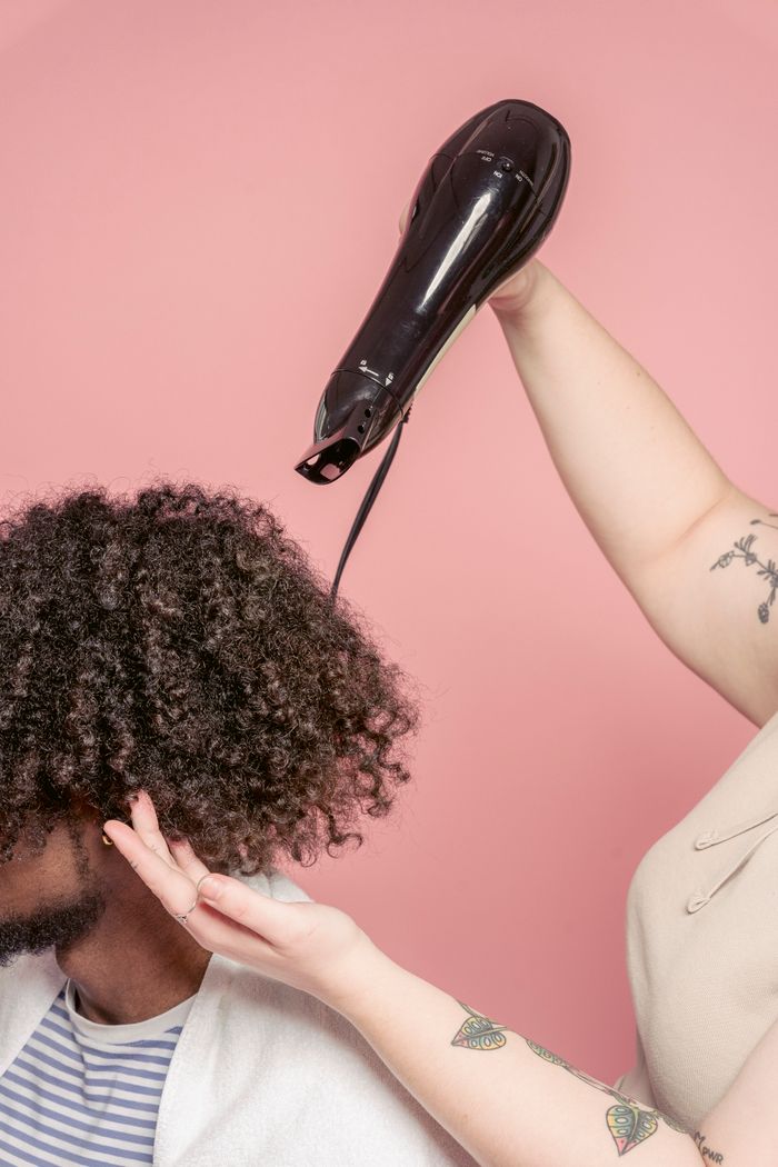 Home Faceless female hairstylist with tattoo drying curly hair of unrecognizable African American man with beard using hairdryer in studio salon against pink wall