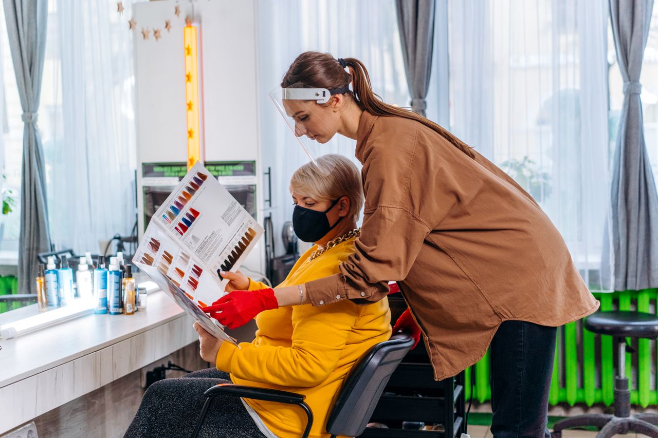 Home Side view female master in medical mask and gloves showing album with hairstyles images to client on chair in salon