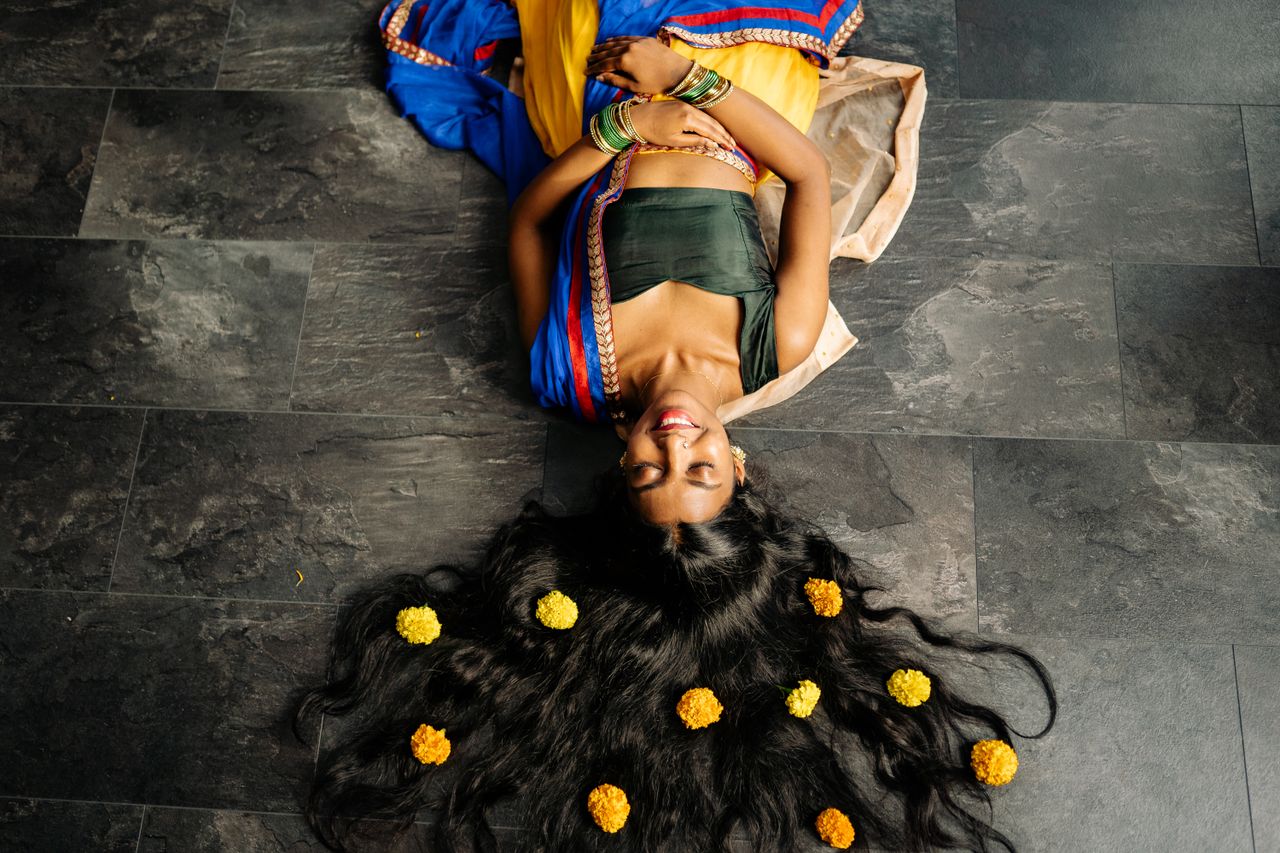 Home A happy Indian woman in traditional attire lies on the floor with flowers woven into her hair.