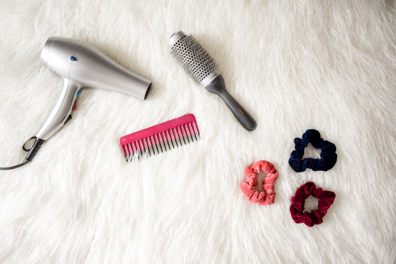 menu-01 Flatlay of hairstyling tools including a hairdryer, brush, comb, and scrunchies on white fur.